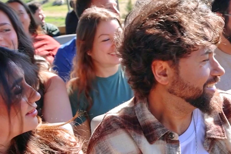 A group of people seated on an outdoor tram, smiling and enjoying a sunny day with a natural landscape in the background.