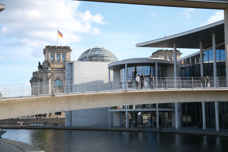 Modern buildings with a bridge over a canal, people standing on the bridge, and a dome with a flag in the background.
