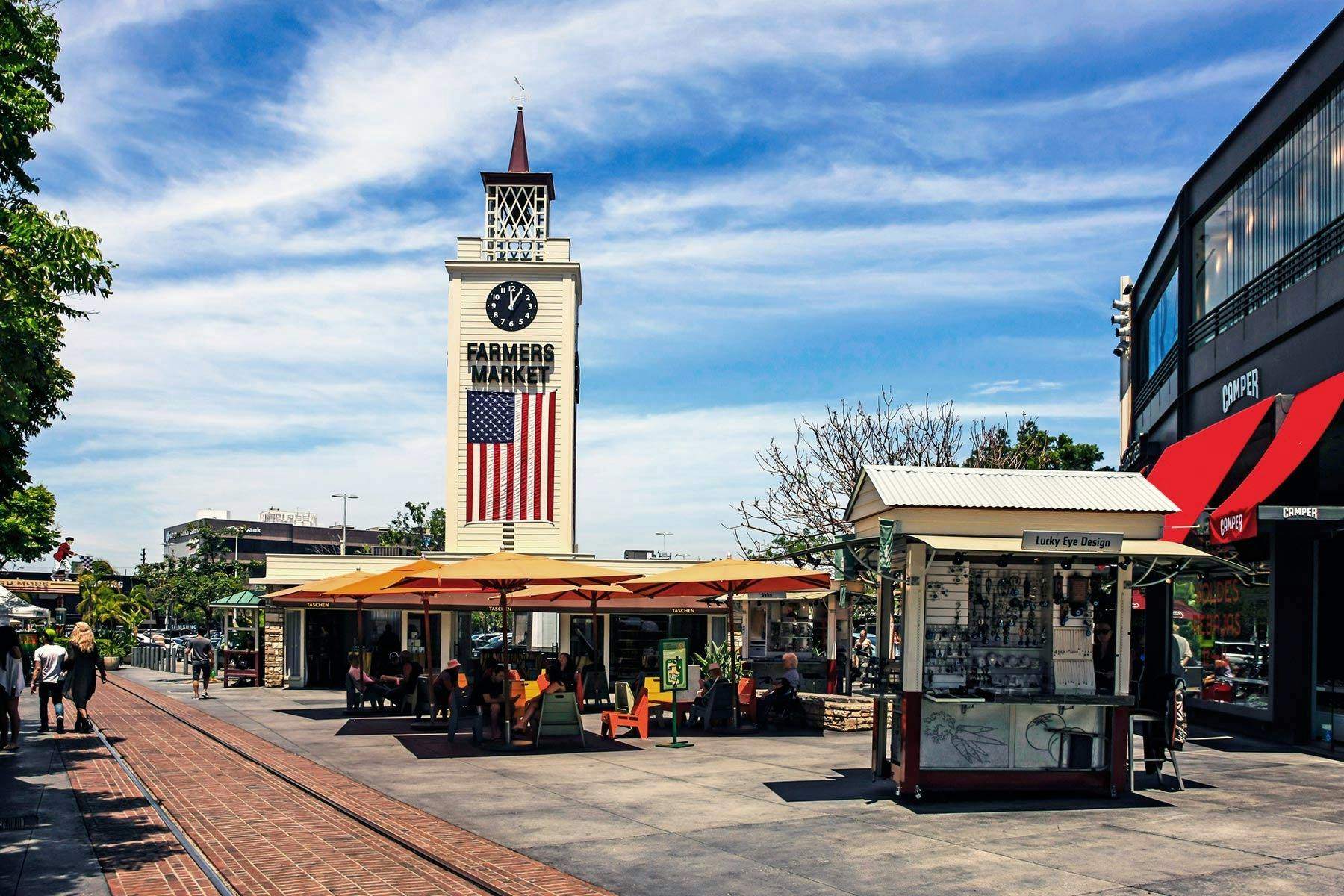 Torre del rellotge en un mercat d'agricultors amb una bandera nord-americana, seients a l'aire lliure sota paraigües i una petita botiga al primer pla.