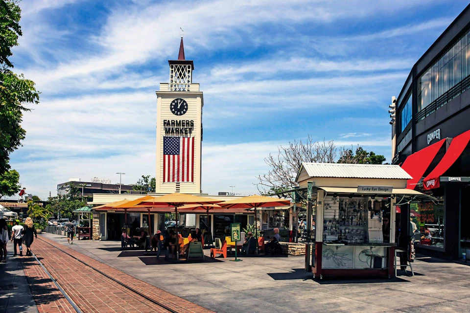 Clock tower at a farmers market with an American flag, outdoor seating under umbrellas, and a small shop in the foreground.