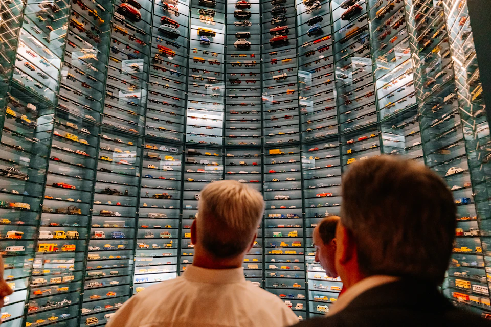 Three people looking at a large, curved wall filled with rows of model cars displayed in glass cases under bright lighting.