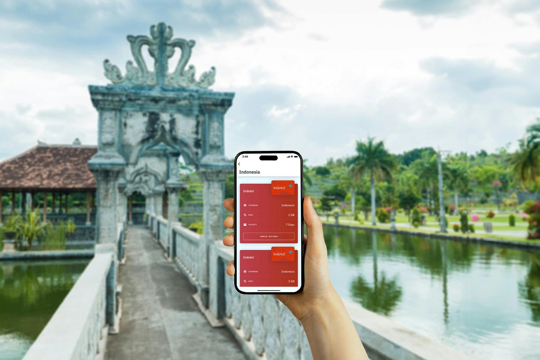 Ruka drží chytrý telefon s indonéskou aplikací eSIM. Pozadí: kamenný obloukový most, voda a tropická zeleň.