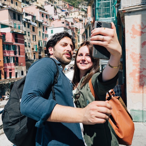Una coppia scatta un selfie, sorridendo di fronte a edifici colorati in una giornata di sole. Entrambi hanno uno zaino e la donna tiene in mano uno smartphone.