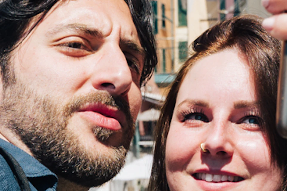 A couple takes a selfie, smiling in front of colorful buildings on a sunny day. They both have backpacks and the woman is holding a smartphone.