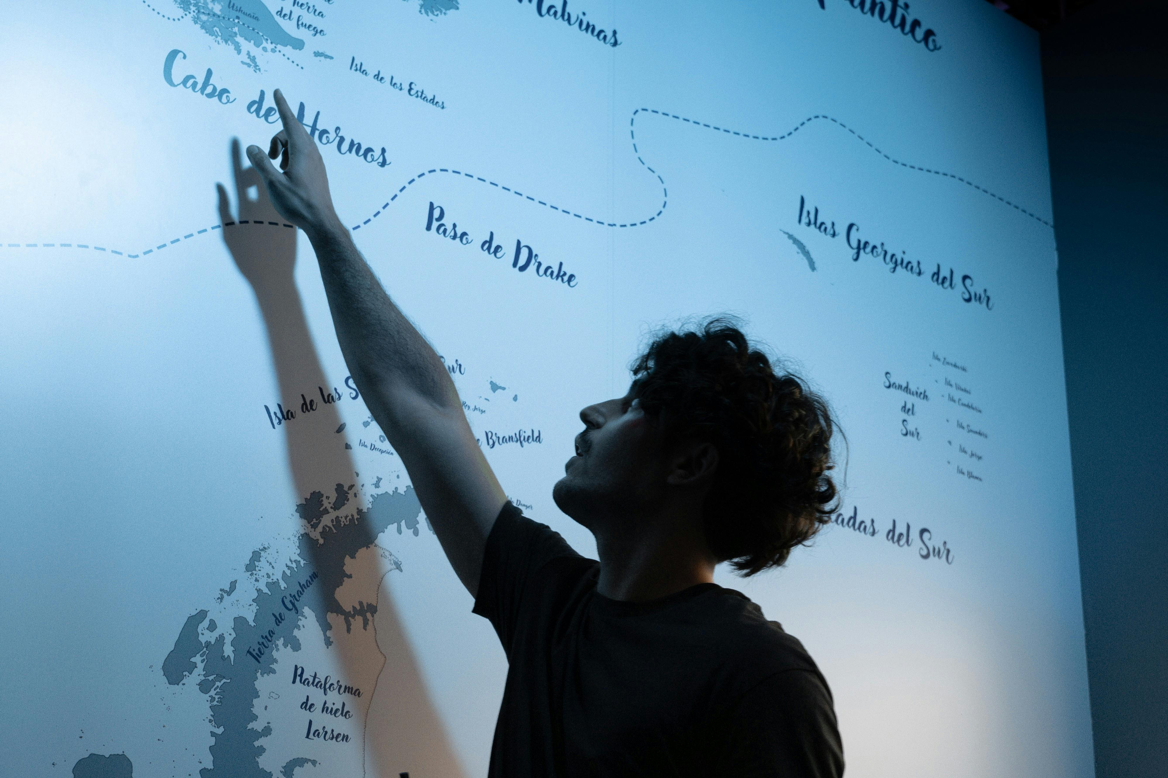 Osoba ukazující na velkou nástěnnou mapu, osvětlenou jemným modrým osvětlením. Mapa zobrazuje různá místa a námořní trasy.