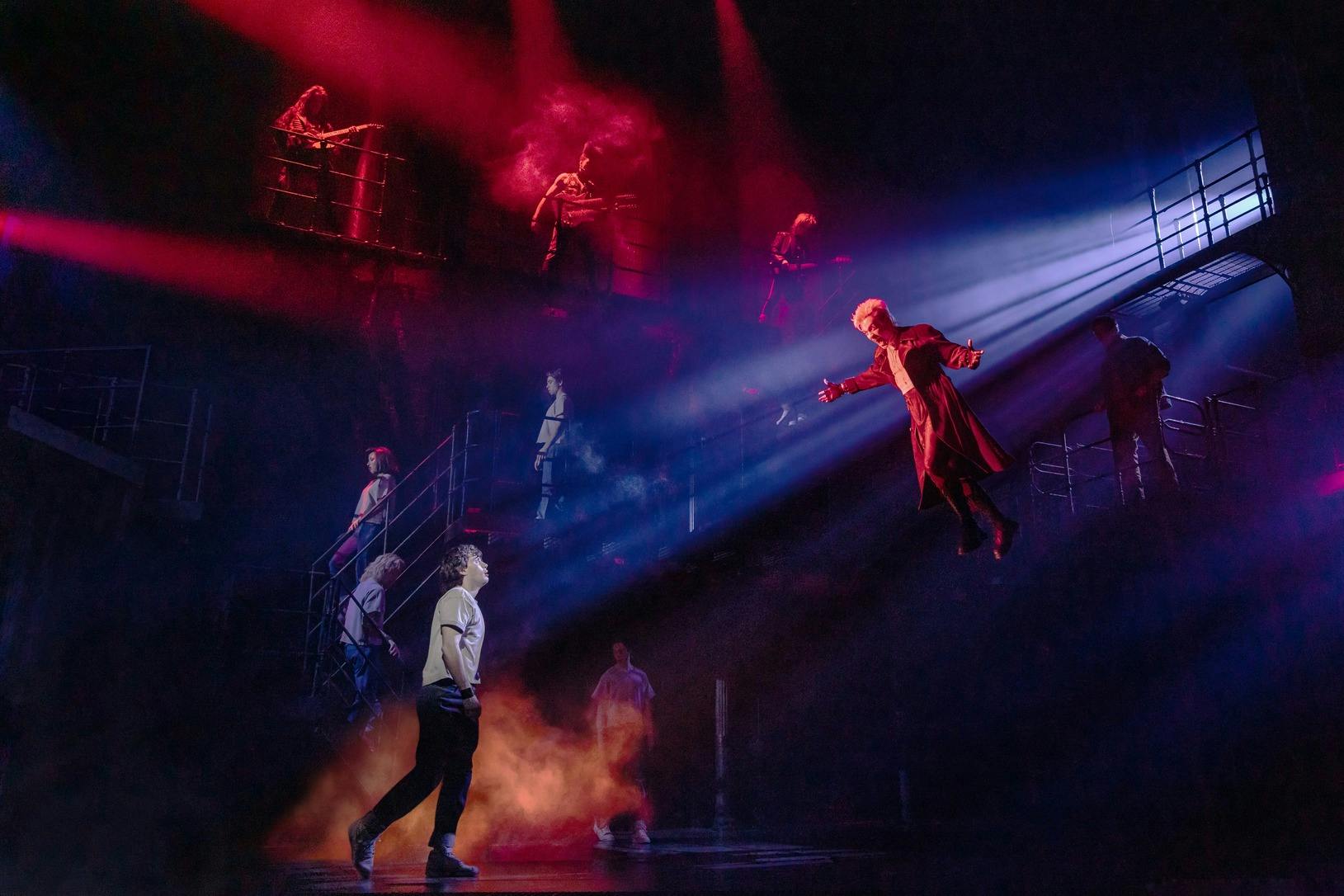 A dramatic stage performance with a person flying under spotlight, surrounded by actors and red lighting effects.