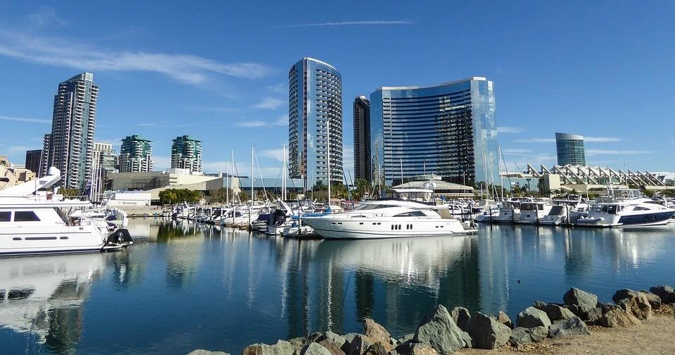 A marina with yachts docked in front of modern high-rise buildings under a clear blue sky.