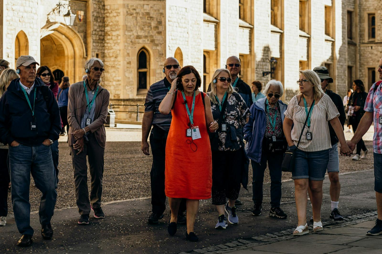 A group of people wearing lanyards and earbuds, walking near a historic stone building with arched windows and doorways.