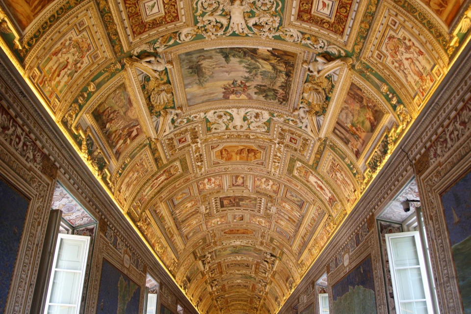 Ornate hall with a richly decorated, gold-accented arched ceiling, featuring intricate paintings and detailed carvings.