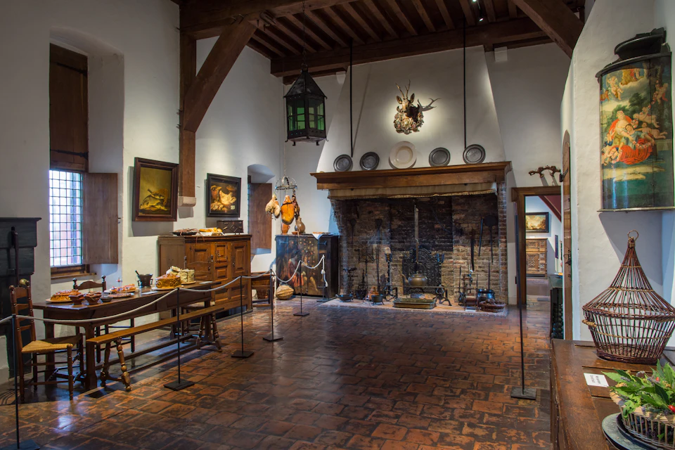 A rustic room with a brick fireplace, wooden beams, paintings and antique furniture, all beneath a high ceiling.