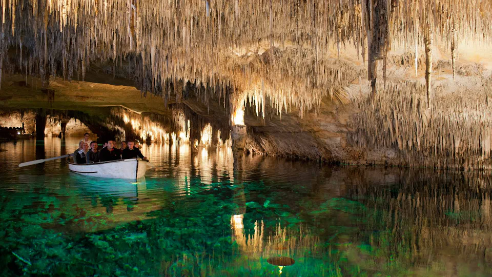 People in a small boat glide through a cave with illuminated stalactites and clear, greenish water.