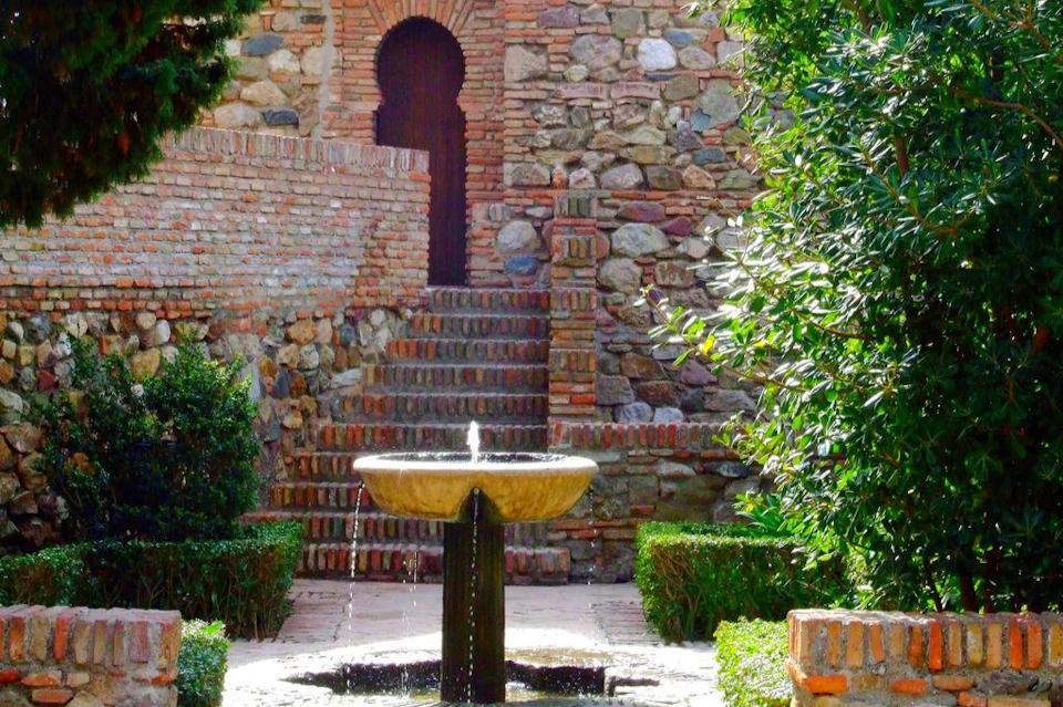 Stone courtyard with a small fountain centered, stone steps leading up to an arched doorway, and greenery on both sides.