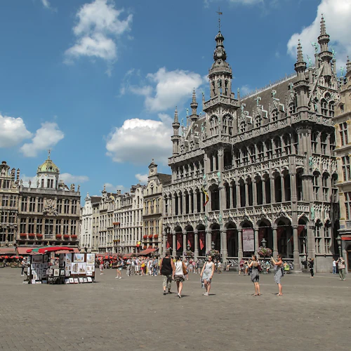 Människor som går runt på ett historiskt torg med utsmyckade byggnader, konstutställningar och röda markiser under en blå himmel med moln.