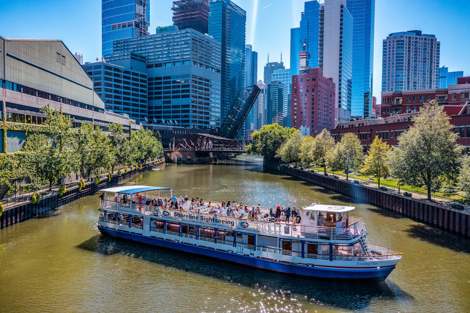 Tour boat with "Shoreline Sightseeing" signage travels along a river, surrounded by tall city buildings and a raised drawbridge.