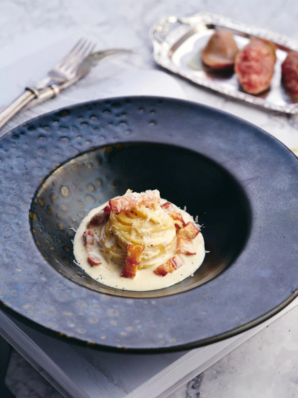 A plate of creamy pasta garnished with bacon bits, served in a dark dish, with a fork and background food visible.
