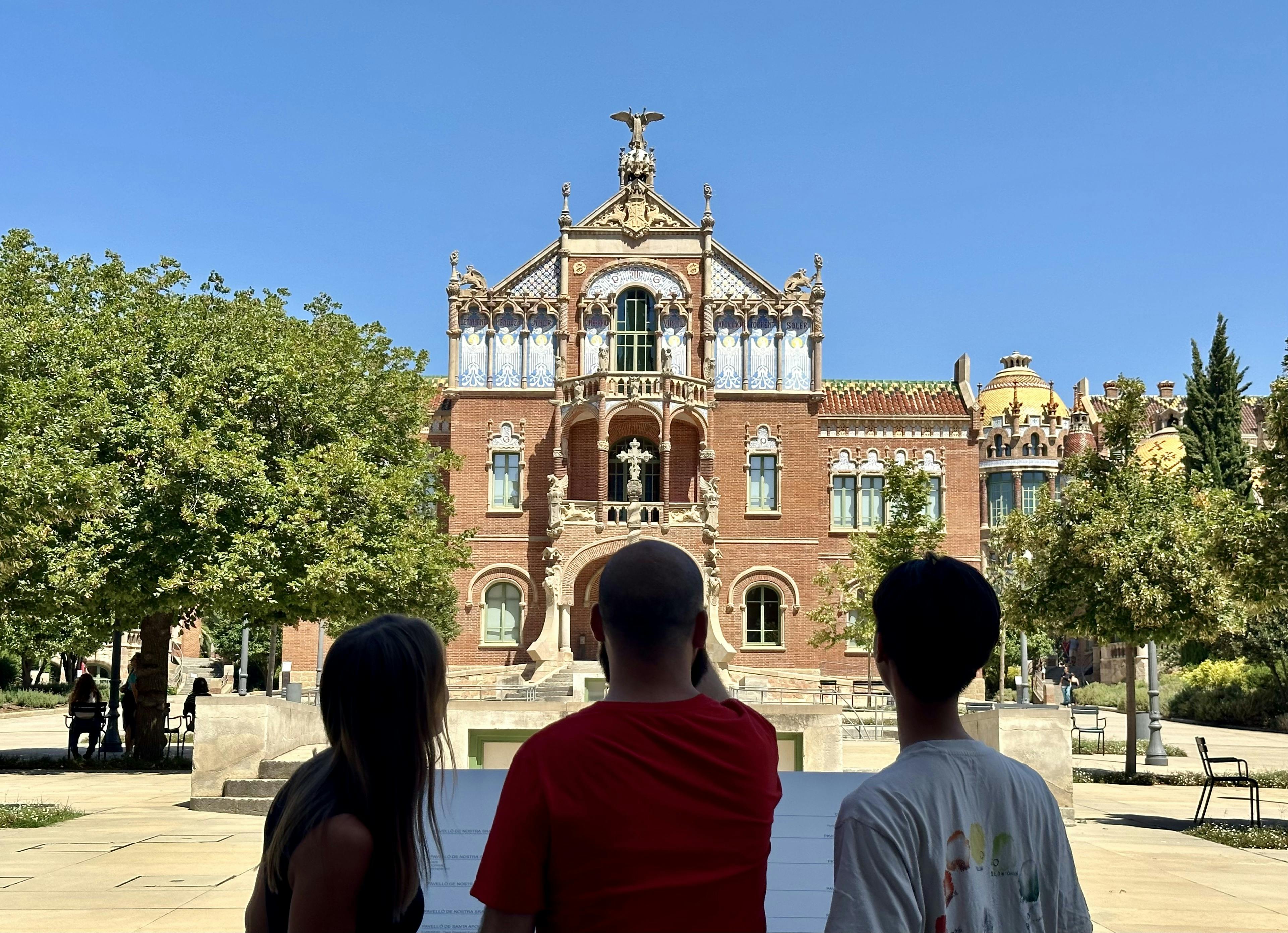 Three people stand looking at an ornate, historical brick building with intricate architectural details under a clear blue sky.