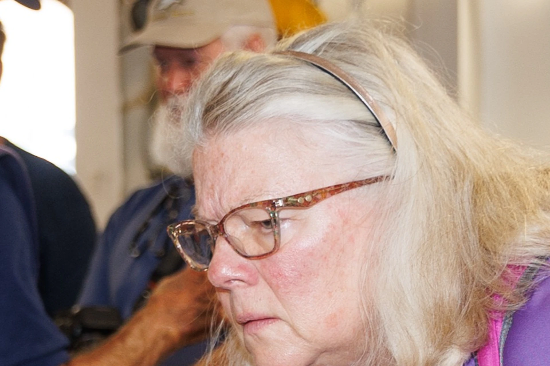 A woman in a purple shirt examines a dining table set with plates, glasses, and a centerpiece, with others in the background.