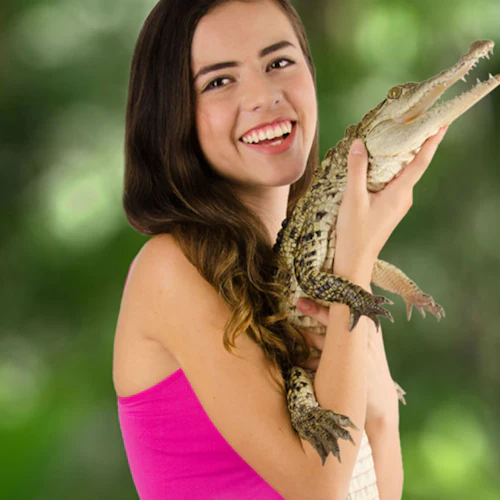 Lächelnde Person in einem rosa Oberteil hält ein kleines Krokodil vor einem unscharfen grünen Hintergrund.