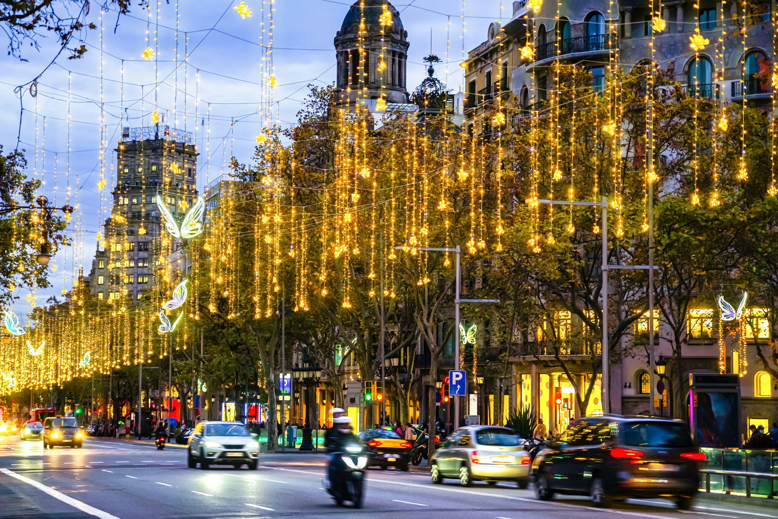 A busy city street at dusk, adorned with golden hanging lights and butterfly decorations, with cars and motorcycles passing by.