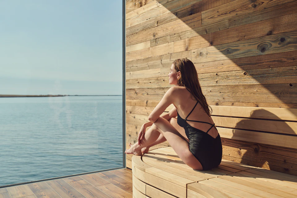 A woman in a swimsuit sits on a wooden bench inside a sauna, looking out at a serene water view through a large window.