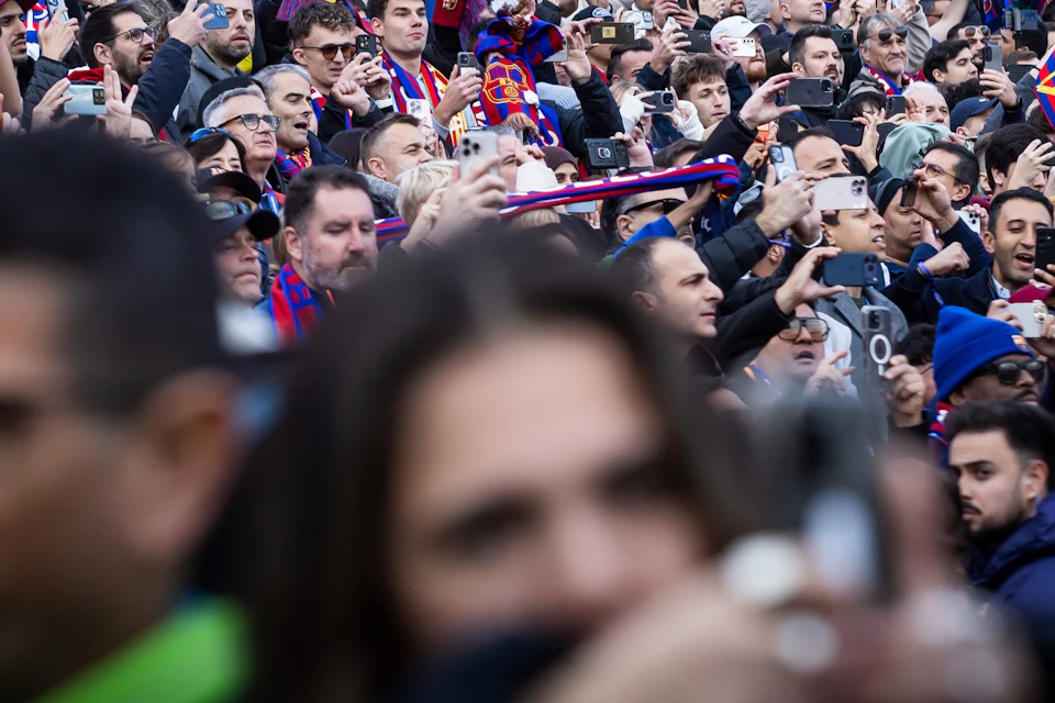 A crowd of sports fans in a stadium, many holding scarves with the text "FC Barcelona," under a large screen displaying "EL SA."