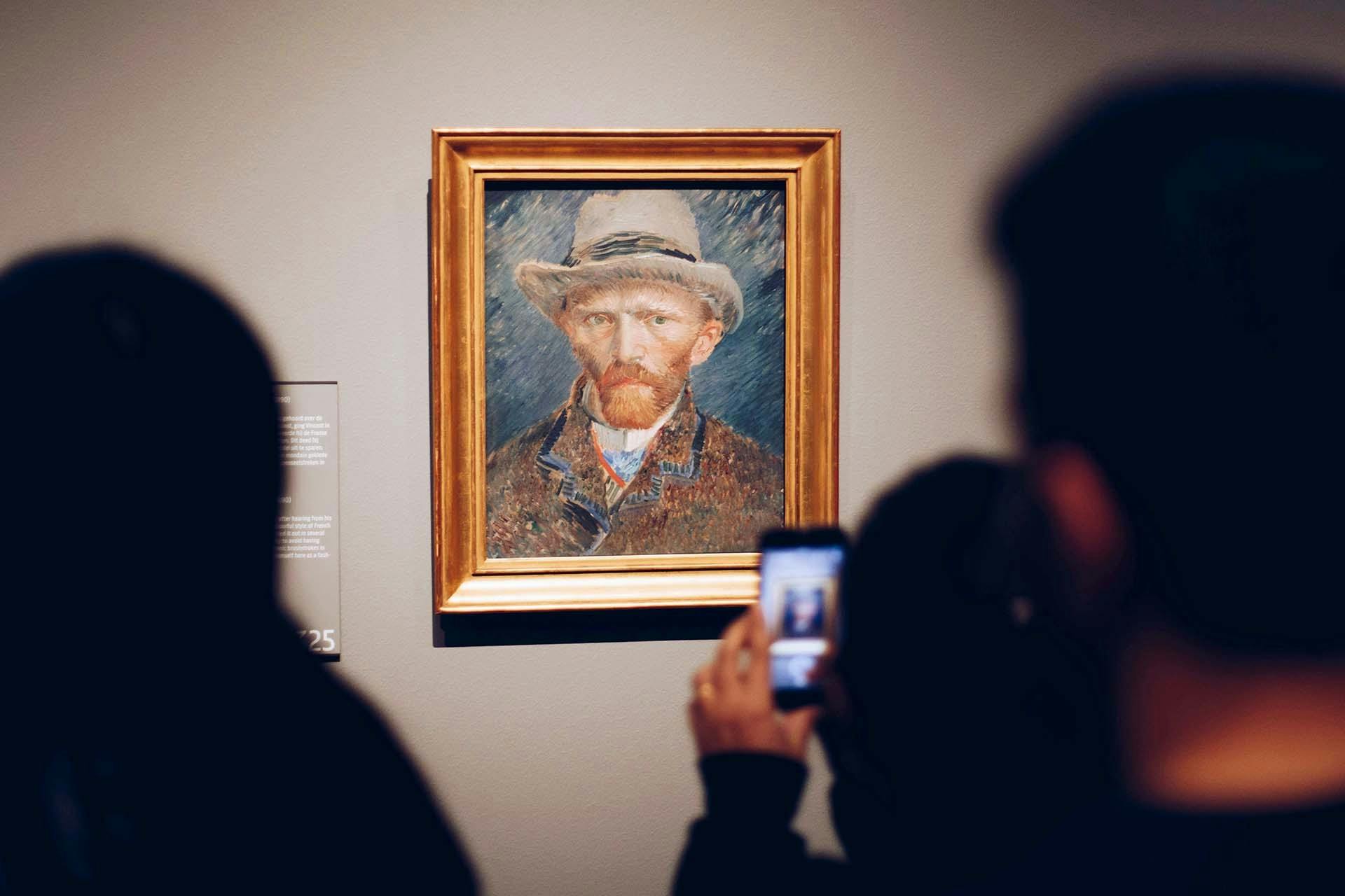 Uma pintura emoldurada de um homem com um chapéu está exposta numa parede, com os espectadores a fotografarem-na em primeiro plano.