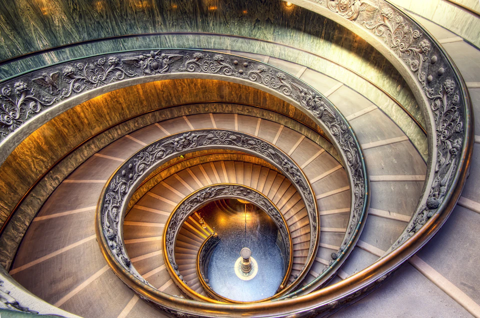 Intricately designed spiral staircase with ornate railings, winding downwards and illuminated by warm lighting.