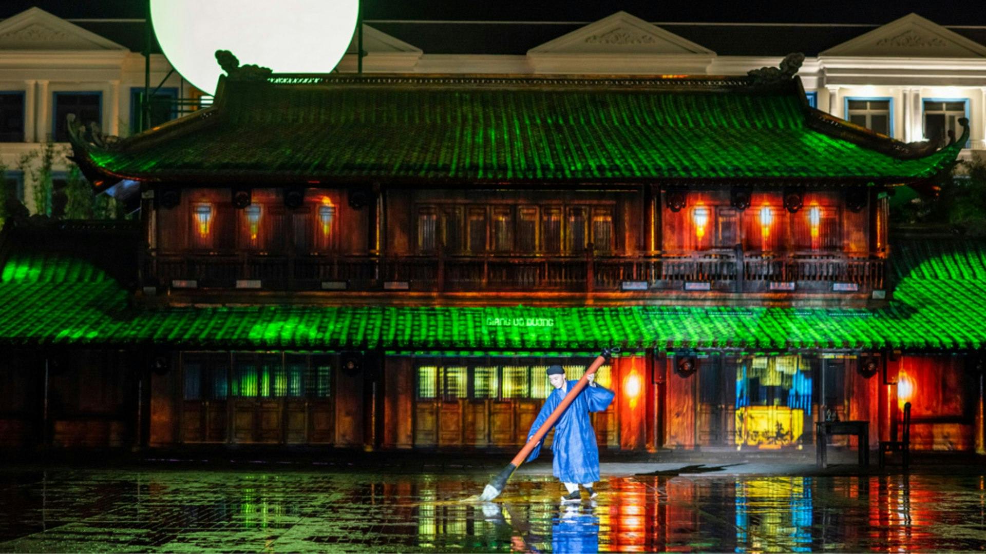 A person in a blue outfit holding a large brush stands in front of an illuminated building with green and orange lighting.