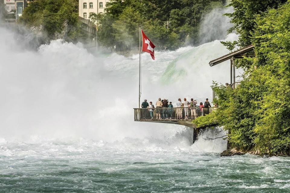 People standing on a platform by a waterfall with a Swiss flag waving, surrounded by mist and lush greenery.