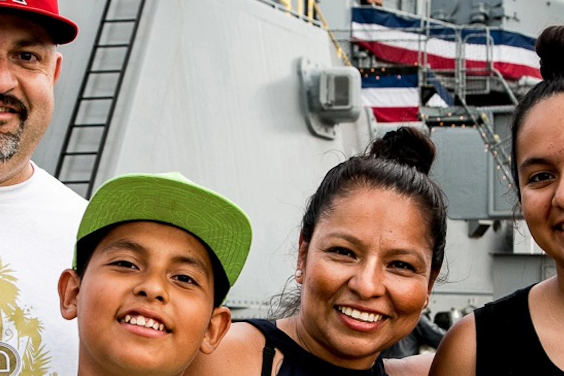 A smiling family of four poses in front of a large naval ship with patriotic decorations, giving thumbs-up gestures.