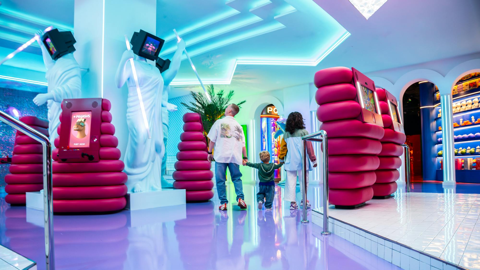 Rodina prochází barevným futuristickým pokojem s neonovými světly, pixelovými sochami a plyšově růžovým posezením.