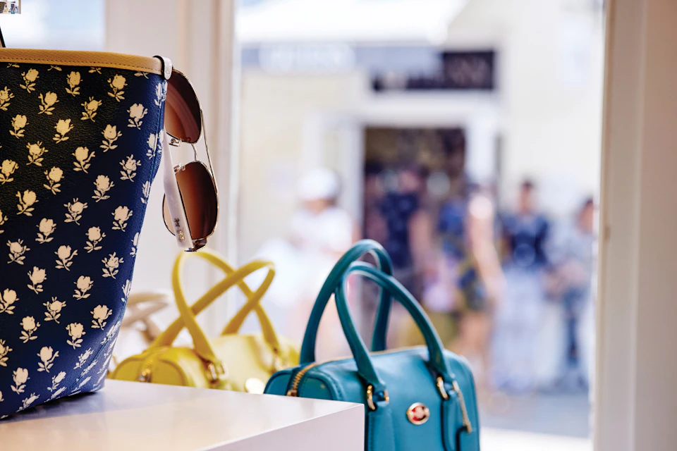 A display of handbags and sunglasses in a store, with blurred people walking outside in the background.