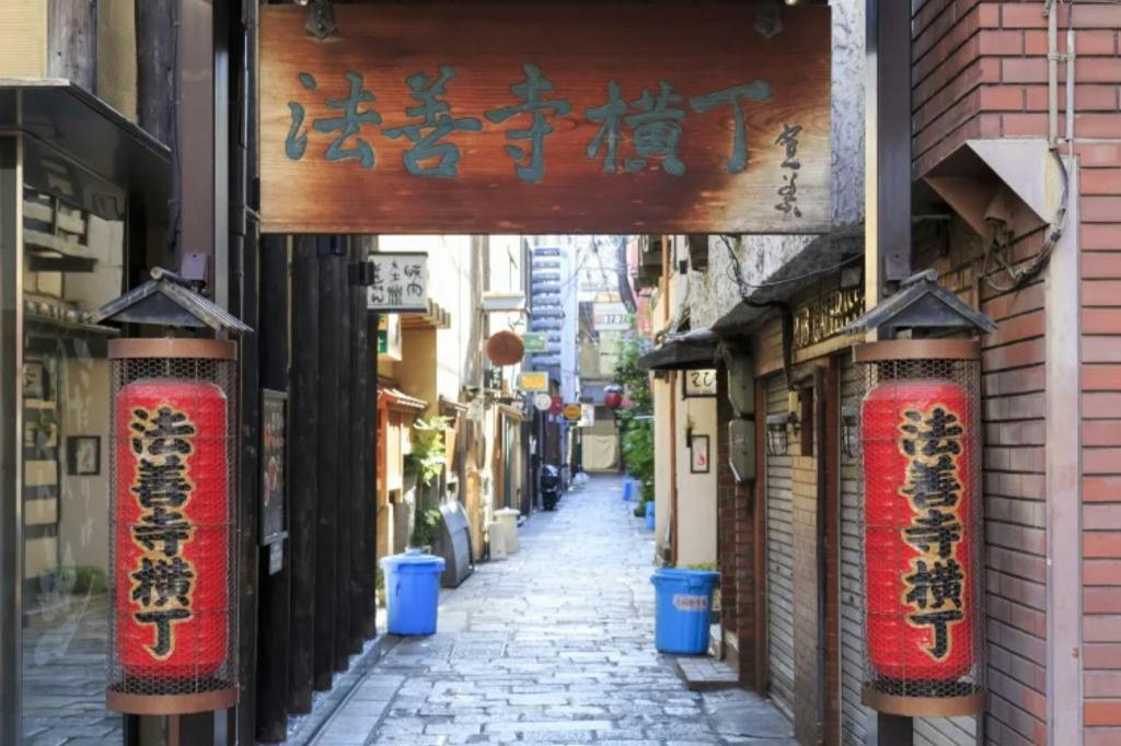 A narrow, paved alley lined with small buildings, red lanterns, and a wooden sign with Japanese characters at the entrance.
