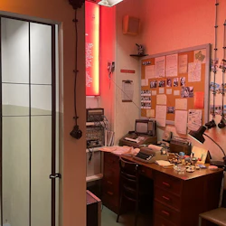 Ett vintagekontor med skrivmaskin, liten TV och pappersarbete på ett skrivbord och en cell med spärrad dörr till vänster. Anslagstavla med foton och dokument.