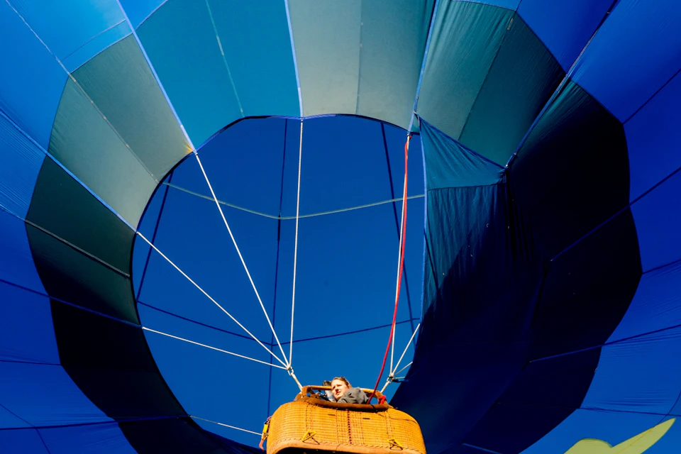 Person standing in a hot air balloon basket, looking up at the large blue and teal balloon against a clear blue sky.