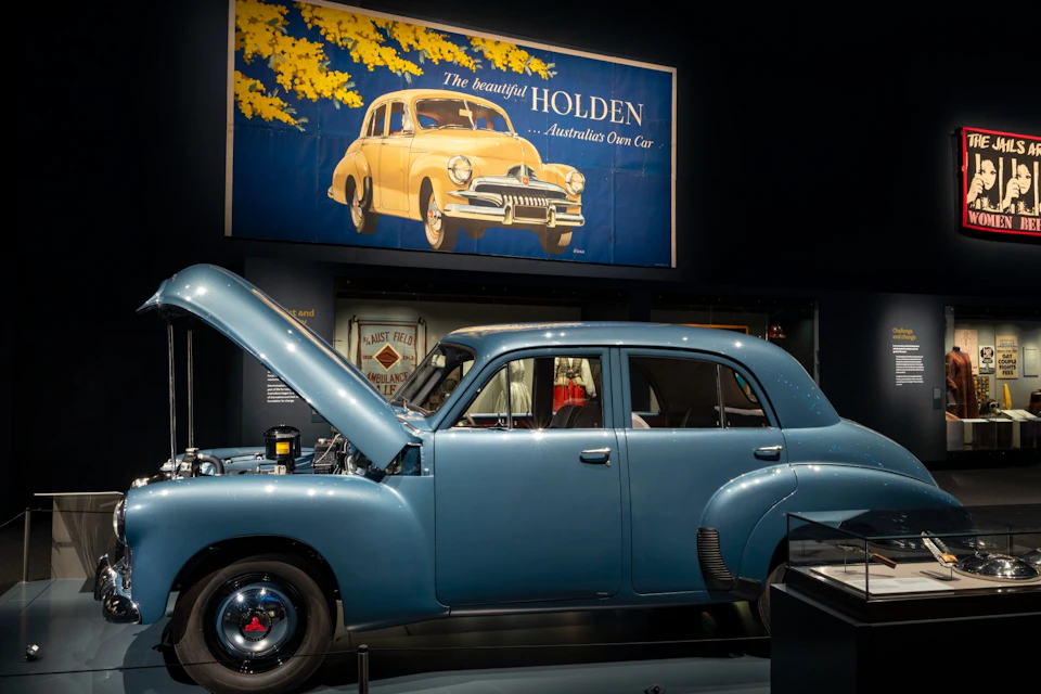 A vintage blue Holden car with its hood open, displayed in a museum setting, featuring a retro Holden advertisement above.