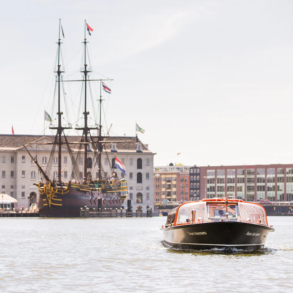 A canal boat approaches, with a historic sailing ship and buildings visible in the background on a sunny day.
