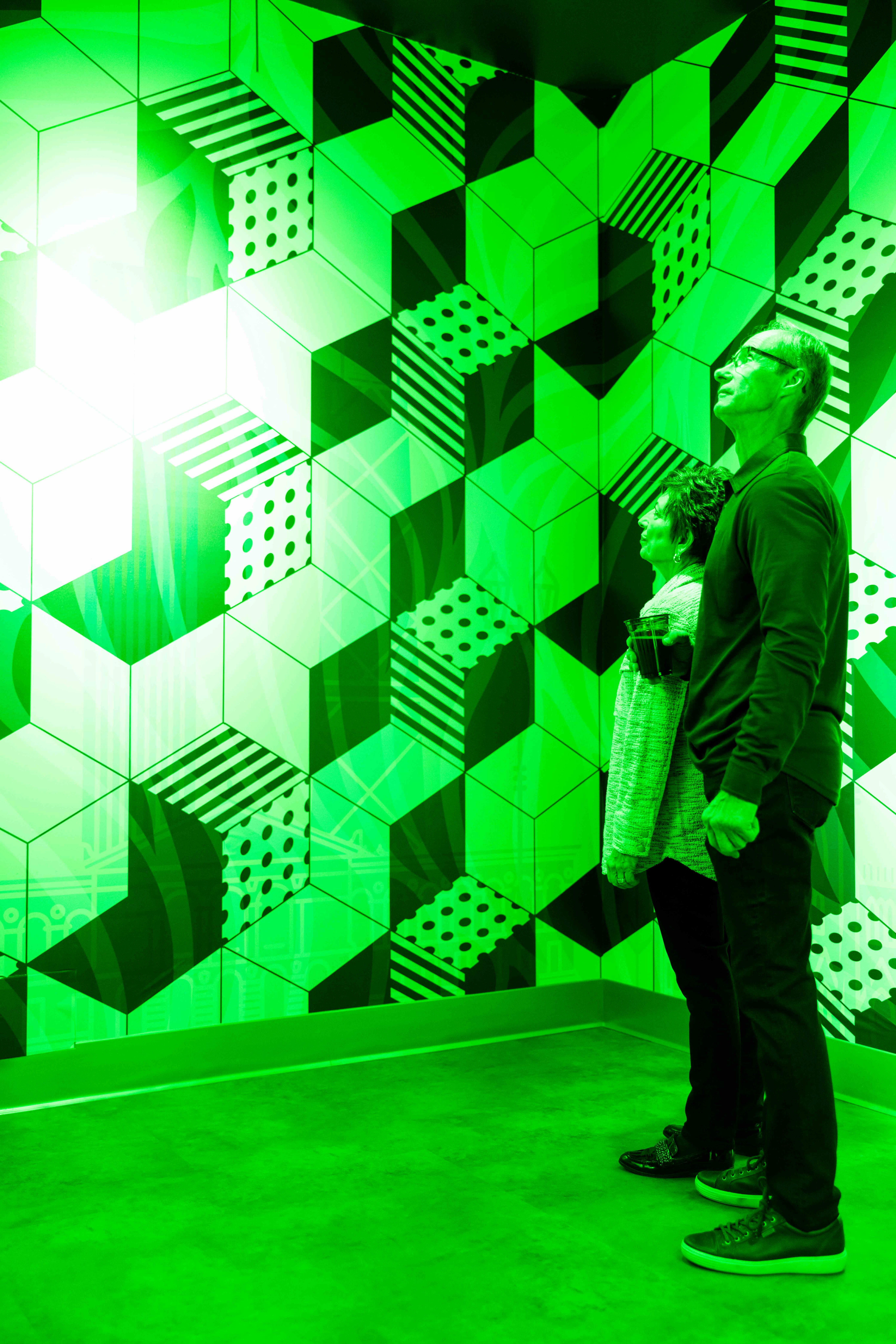 Two people stand in front of a green geometric-patterned wall, gazing upwards towards a bright light.