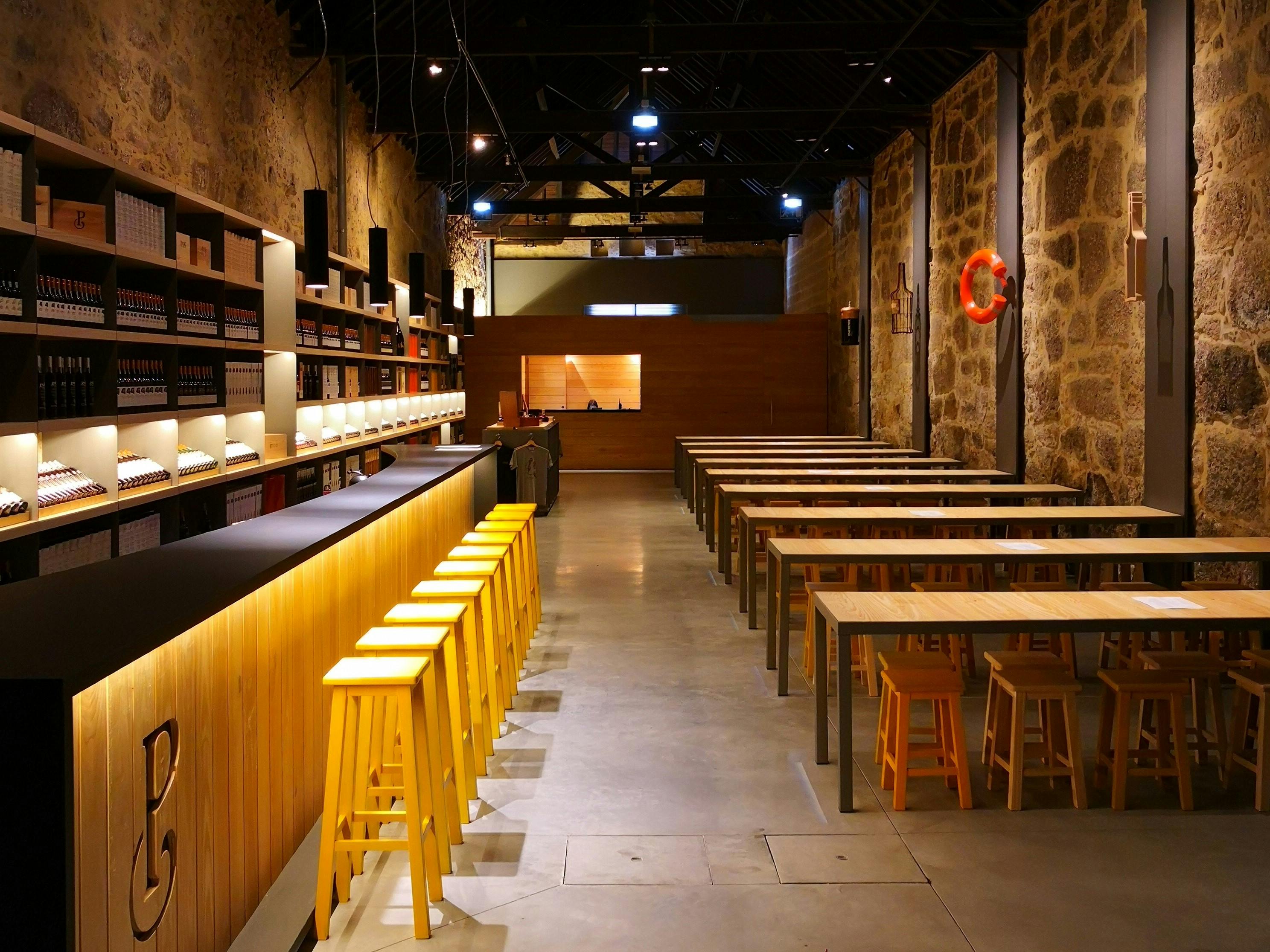 黄色いスツール、長い木製のテーブル、石の壁があるモダンなバー。 ボトルの棚と点灯したバックカウンターが見えます。