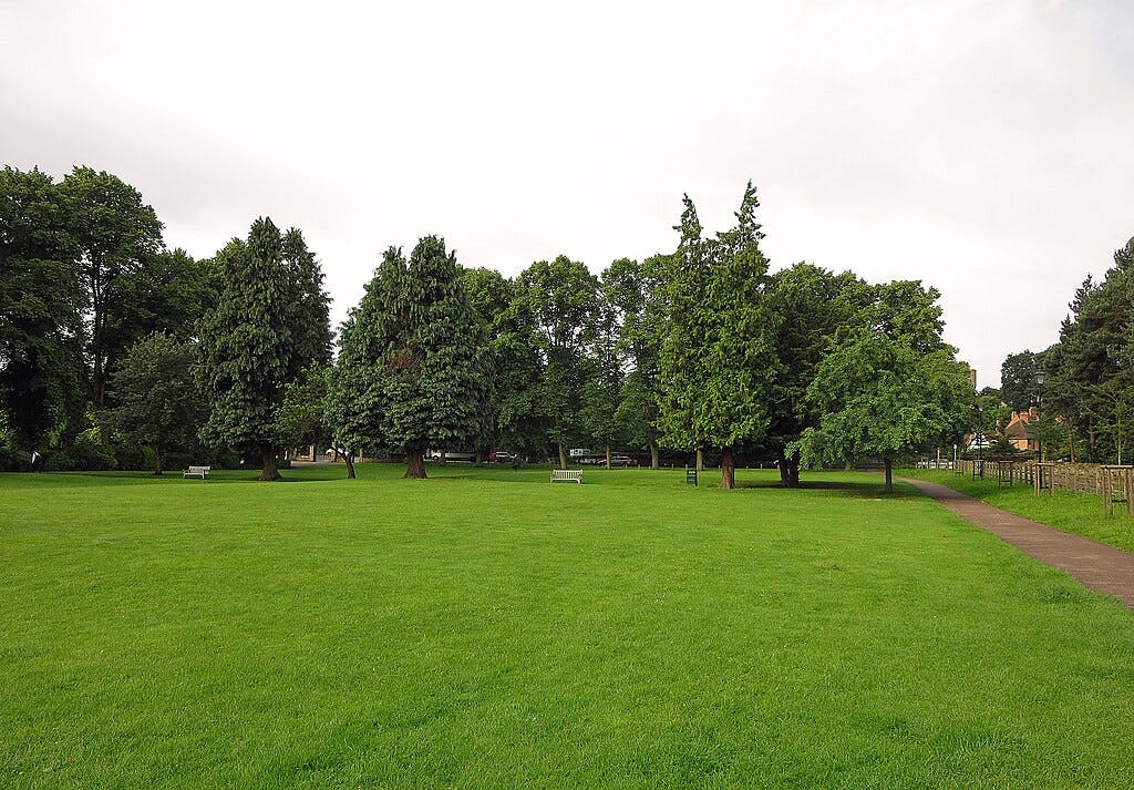 A green park with scattered trees, benches, a paved path, and buildings in the background.
