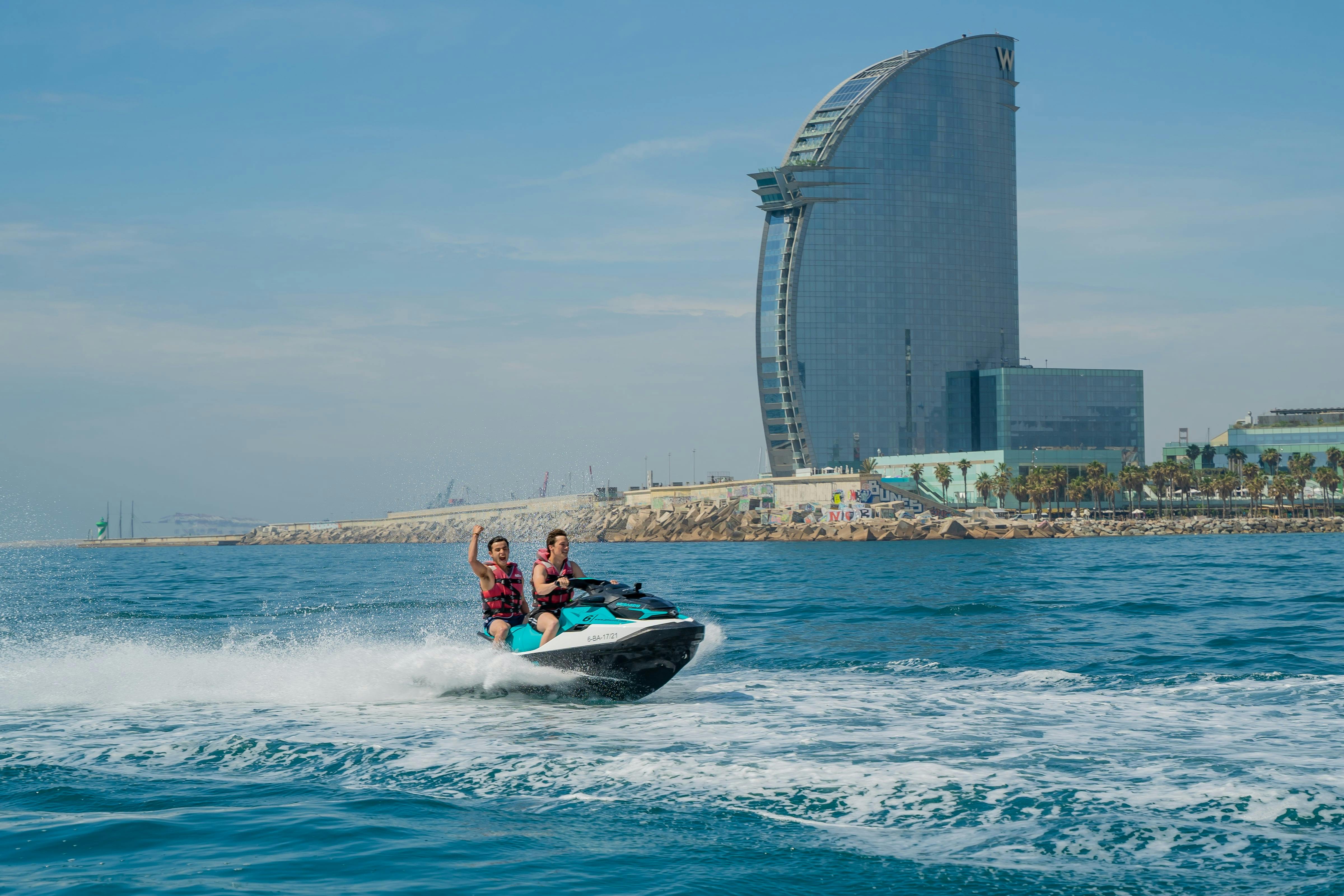 Deux personnes en jet ski sur la mer avec un grand bâtiment hôtelier incurvé et la côte en arrière-plan.