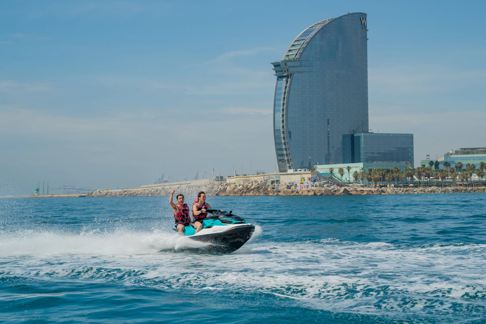 Two people riding a jet ski on the sea with a tall, curved hotel building and coastline in the background.