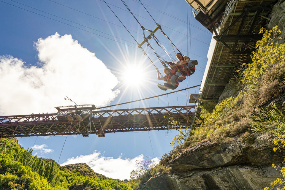 People swinging on a giant swing under a bridge, with sunlight and a scenic backdrop of trees and blue sky.