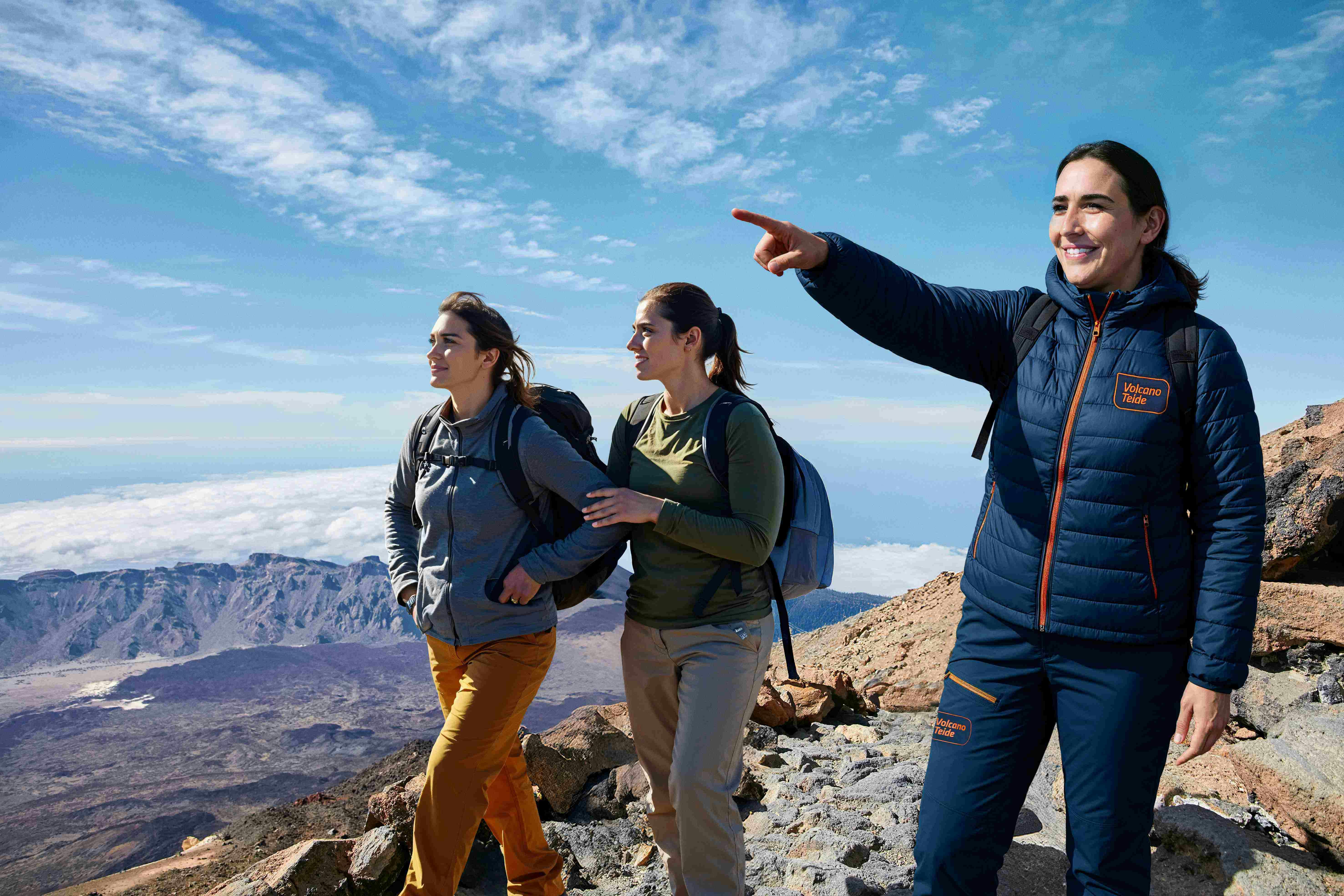 Three women wearing hiking gear stand on a rocky mountain trail; one points and smiles while the other two look in the same direction.