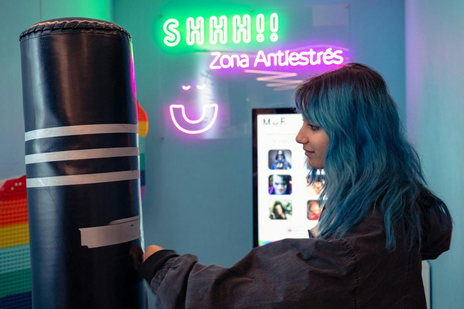 カラフルな部屋の中で、青い髪の人が「シーッ！ ！」と「ゾナ・アンティストレ」と書かれたネオンサインの近くに立っています。