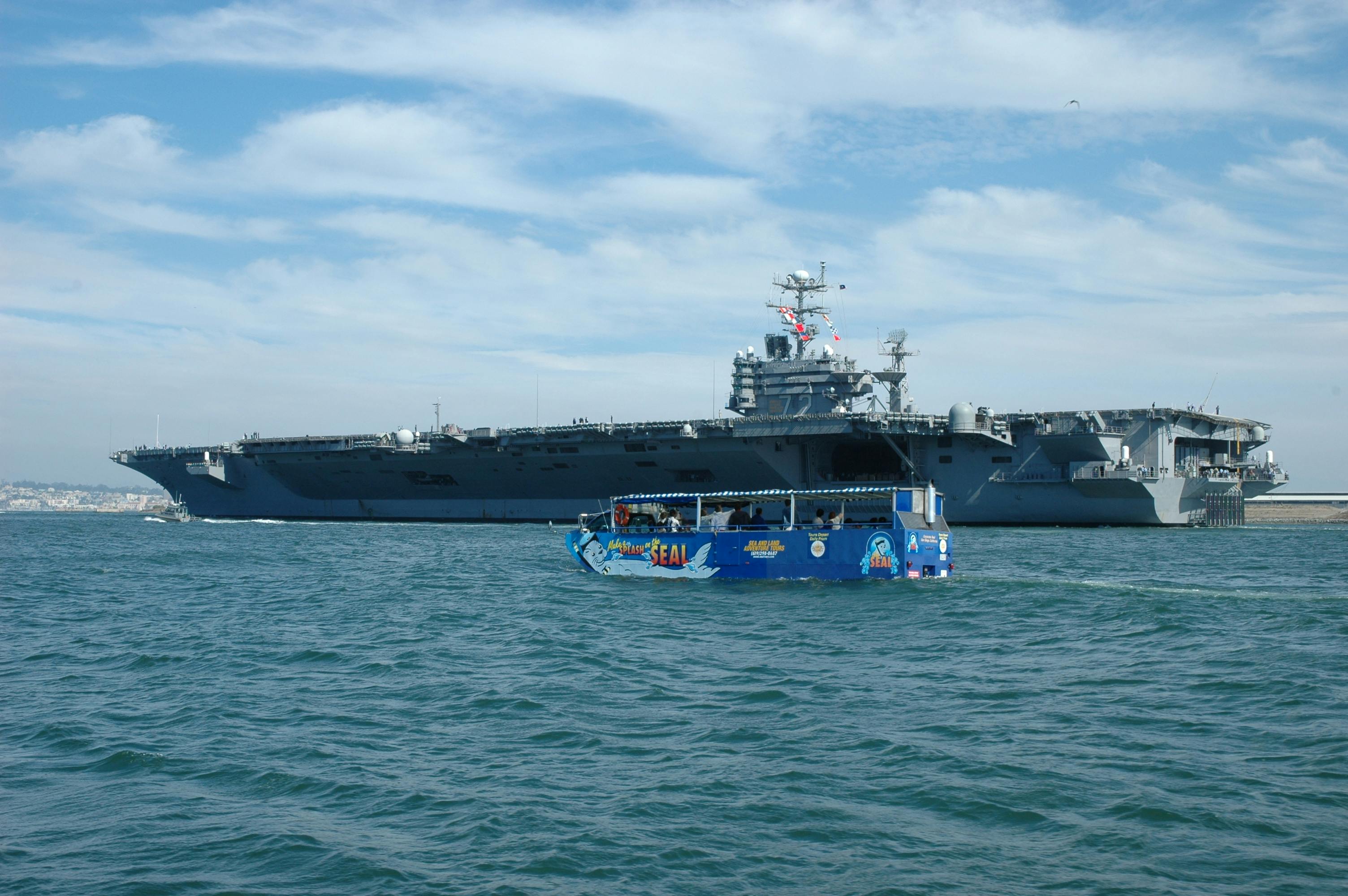 A large aircraft carrier in the background with a colorful blue aquatic-themed tour boat in the foreground on a calm sea.