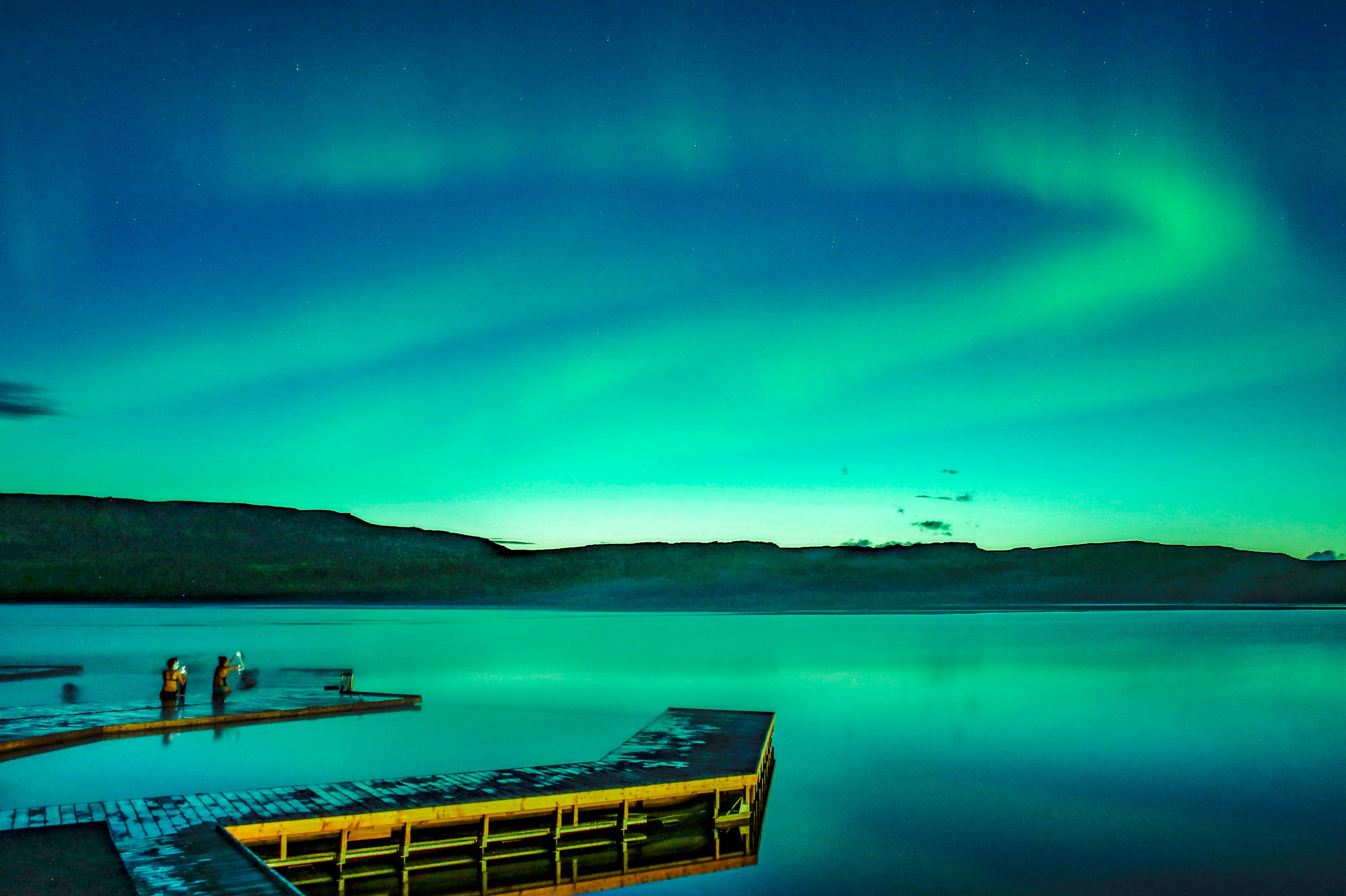 在充满活力的绿色和蓝色北极光下，一个木制码头延伸到一个平静的湖泊中。