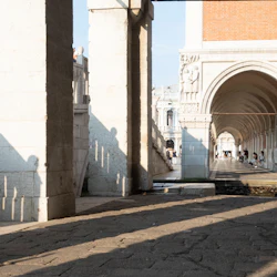 Stenvalv med invecklade sniderier och pelare, skuggad gångväg, människor som sitter och ljus som strömmar in från sidan.