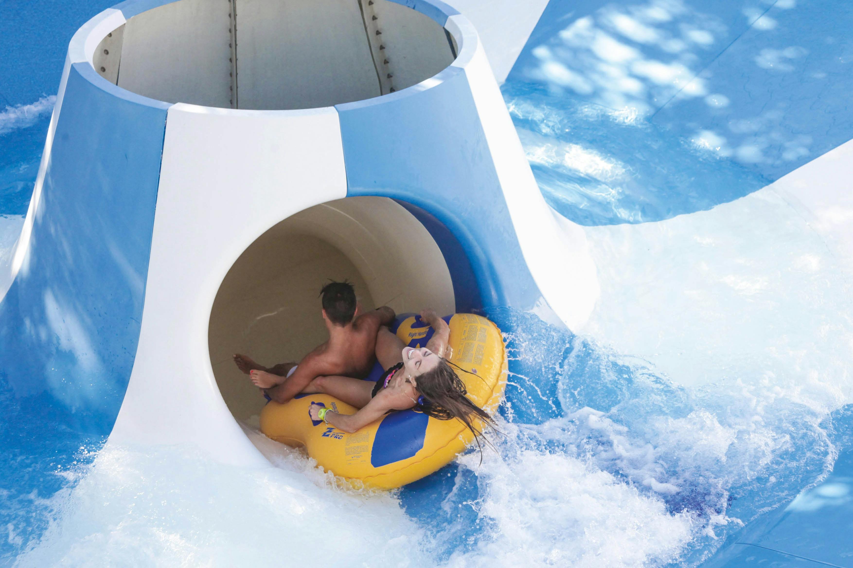 Due persone in sella a un tubo gonfiabile giallo che escono da uno scivolo d'acqua verso una piscina, circondati da spruzzi d'acqua.