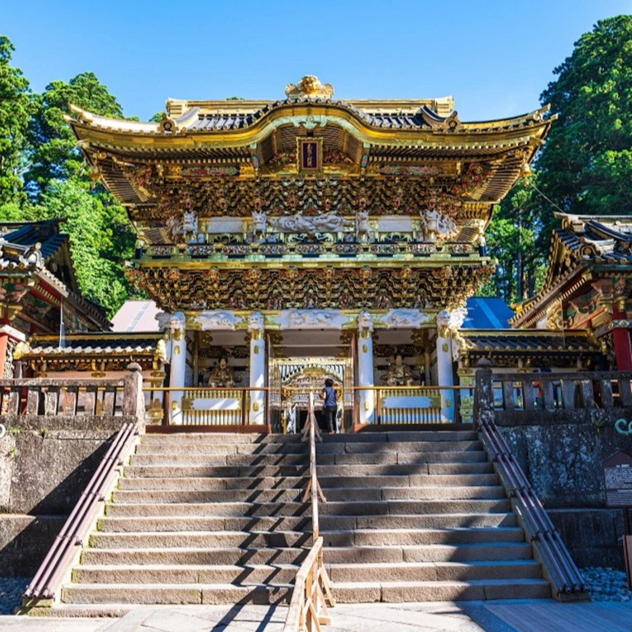 Elaborately decorated golden temple gate with intricate carvings, a staircase leading up, and trees in the background.